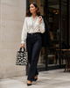 A woman walking outdoors in front of a building with stone walls and glass doors, wearing a cream long-sleeve blouse, dark high-waisted flared jeans, black heels, and carrying a black-and-white patterned tote bag while holding a dark coat.